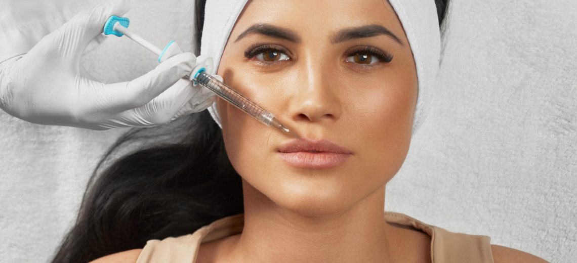 From above view of hyaluronic injection in female lips. Cosmetologist using syringe with liquid while brunette patient in towel on head lying, looking at camera. Concept of cosmetology, beauty.