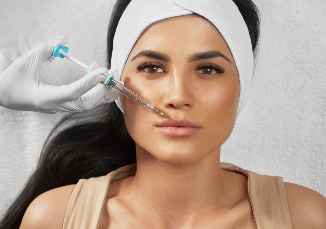 From above view of hyaluronic injection in female lips. Cosmetologist using syringe with liquid while brunette patient in towel on head lying, looking at camera. Concept of cosmetology, beauty.